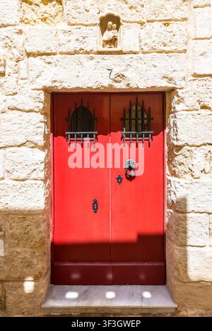 Traditionelle rote Holztür mit schmiedeeisernen Details in einer historischen Kalksteinfassade in der alten befestigten Stadt Mdina, Malta. Architektur Stockfoto