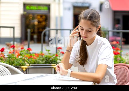 Junge Frau überprüft Smartwatch während eines Telefonats in einem Straßencafé. Junge Frau telefoniert mit einem Smartphone, während sie ihr Smartwatch in einem Straßencafé überprüft Stockfoto