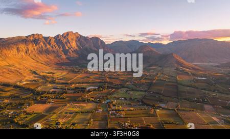 Luftaufnahme des Franschhoek-Tals mit üppigen Weinbergen, sanften Hügeln und dramatischen Bergketten, die vom warmen Schein des Sonnenuntergangs beleuchtet werden Stockfoto