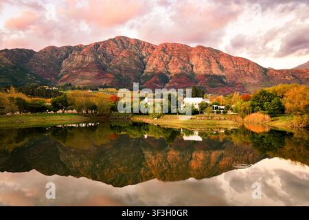 Luftaufnahme der Franschhoek-Berge, die sich in einem ruhigen See spiegeln, umgeben von üppigen Herbstbäumen und Weinbergen während der goldenen Stunde in Franschhoek, We Stockfoto