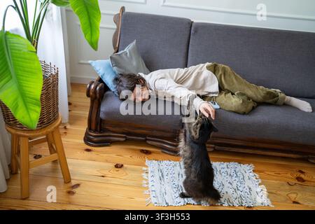 Fröhliche Frau spielt mit Katze, während sie sich vollständig entspannt auf der Couch im Zimmer befindet, Glück zu Hause am Wochenende. Stockfoto