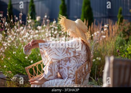 Junge Frau entspannt sich auf einem Stuhl im Sommergarten, trägt ein stilvolles Gewand und trinkt Kaffee. Die Dame trägt einen Strohhut und genießt die Sonne zwischen Blumen Stockfoto