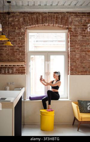 Sportliche Frau sitzt auf der Fensterbank und benutzt ein Smartphone Stockfoto