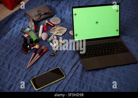 Nahaufnahme eines Laptops, der einen Greenscreen anzeigt, neben einem Smartphone, Kosmetikprodukten und Hautpflegewerkzeugen auf einem Bett, was die Erstellung von Beauty-Inhalten nahelegt. Nützlich für E-Commerce, Influencer-Marketing, Mockup Stockfoto