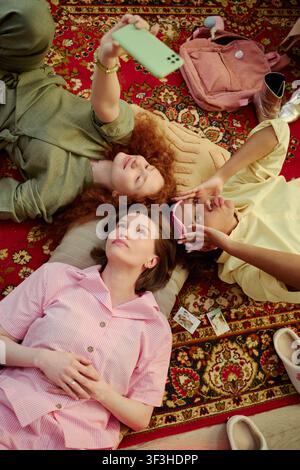 Vertikale Aufnahme von verschiedenen jungen erwachsenen Frauen, die auf einem Teppich liegen, ein Smartphone-Selfie machen und zusammen entspannen. Repräsentiert Freundschaft, Social-Media-Inhalte, Jugendlebensstil-Marketing, vertikal Stockfoto