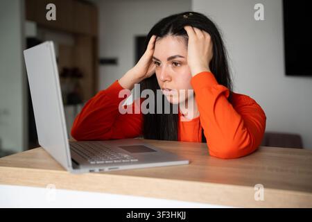 Frustrierte junge Frau, die ihren Kopf hält, während sie auf einen Laptop-Bildschirm drinnen schaut. Stressmanagement digitaler Burnout und Arbeitsplatzherausforderungen. Stockfoto