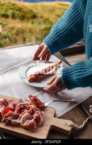 Nahaufnahme von Scheiben gekürten Schinkens, die über einen knusprigen Brotstab drapiert sind, gehalten von weiblichen Händen. Informelle Verkostungsszene in rustikalem Kontext. Schinken, der mit Stockfoto