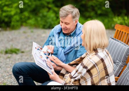 Liebevolles Seniorenpaar sitzt in Liegestühlen und bewundert Fotos in Familienalbum auf dem Land Stockfoto