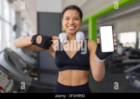 Lächelnde junge schwarze Frau, die in einem Fitnessstudio auf ein Smartphone mit leerem Bildschirm zeigt Stockfoto