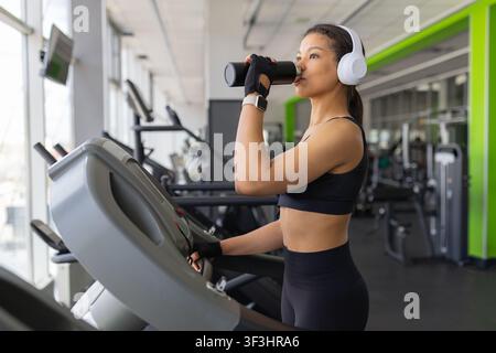 Schwarze Frau trinkt Wasser aus einer wiederverwendbaren Flasche, während sie auf dem Laufband im Fitnessstudio unterwegs ist Stockfoto