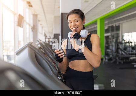 Lächelnde schwarze Frau isst Proteinriegel und benutzt ihr Smartphone im Fitnessstudio Stockfoto