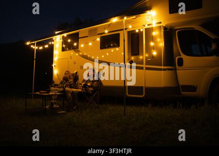 Paar sitzt an einem Tisch vor ihrem Wohnmobil und genießt einen gemütlichen Abend unter warmem Lichterkettenlicht, verbringt Qualitätszeit zusammen während eines Stockfoto