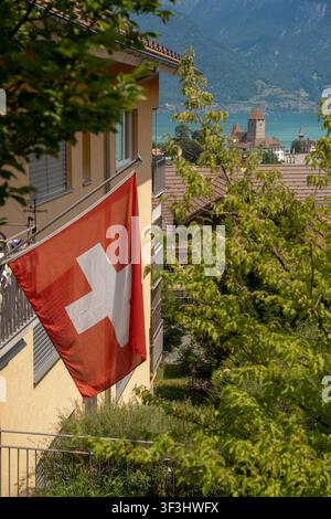 Spiez, Schweiz – 28. Juni 2026: Helle Tagesansicht einer Schweizer Nationalflagge, die von einem Wohnbalkon in Spiez, Schweiz, hängt und überblickt Stockfoto