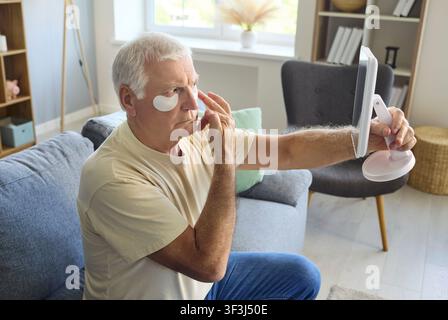 Senior applying unter Augentupfer mit Spiegel zu Hause, Hautpflegeroutine Stockfoto