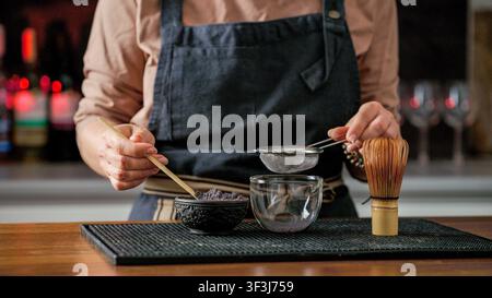 Person, die eine Schürze trägt, verwendet einen Bambislöffel, um violettes Teepulver in eine Glas Tasse in einem Café zu sieben Stockfoto