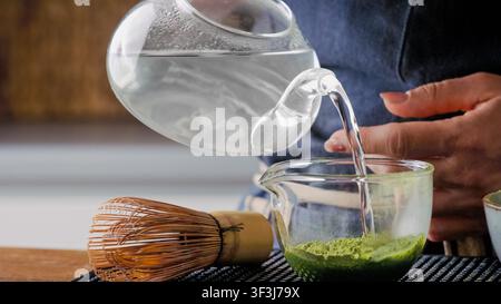 Heißes Wasser aus einer Glas-Teekanne in eine Schüssel mit Matcha-Pulver und Bambus-Rührer auf dem Tisch gießen Stockfoto