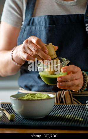 Frau, die grünes Matcha-Pulver in einer Glasschale auf einem Holztisch während der Morgenvorbereitung aufschlägt Stockfoto