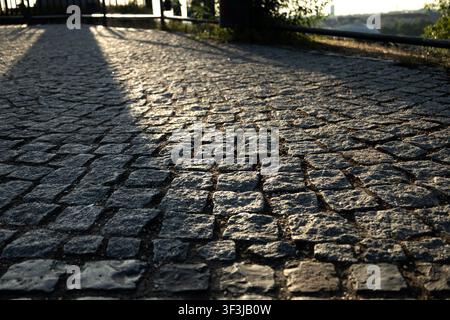 Eine niedrige Ansicht einer leeren Kopfsteinpflasteroberfläche mit warmem Sonnenlicht und langen Schatten. Die raue Steinstruktur füllt das Bild mit einer geringen Tiefenschärfe Stockfoto