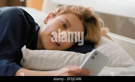 Lächelnde kaukasische Frau, die auf einem Handy scrollt, ruht, entspannt, liegt in einem gemütlichen Bett am Morgen, positives Mädchen, lächelt, durchstöbert Smartphone-App-Web Stockfoto