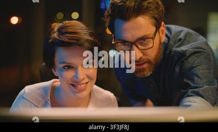 Kaukasischer Geschäftsmann schaut auf Laptop im Abendbüro, prüft Projektergebnisse, Frau lächelt, weibliche Zuhörerin, Firmen-CEO hilft mit dem Computer, Unternehmensumgebung Stockfoto
