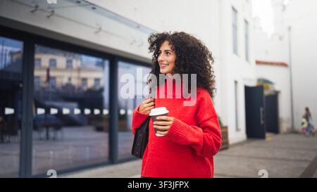 Lockenköpfige kaukasische Frau in rotem Pullover, die eine Tasche trägt und einen Takeaway-Kaffeebecher hält, während sie draußen in der Nähe einer weißen Wand und von Grünpflanzen geht. Lächelnd Stockfoto