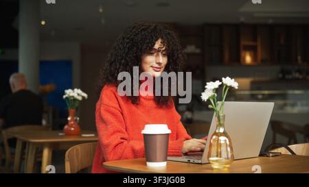 Schöne kaukasische junge Frau mit lockigem Haar, die im Freien in einem Café an einem Laptop arbeitet. Freiberuflerin sitzt an einem Holztisch mit Kaffeetasse und Stockfoto