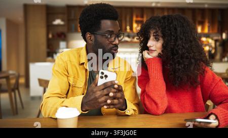 Mann in gelbem Hemd zeigt Smartphone einer Frau in rotem Pullover am Cafétisch. Afroamerikanischer Mann mit Brille hält Telefon, während lockighaarige Stockfoto