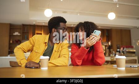 Mann mit gemischter Herkunft in gelbem Hemd und Frau in rotem Pullover sitzen an einem Café-Tisch mit Kaffeetassen und schauen gemeinsam auf ein Smartphone. Mann mit Brille Stockfoto