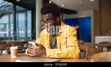 Aufgeregter afroamerikanischer Mann mit Brille lacht, während er im Café sein Smartphone überprüft. Mann lächelt vor Freude, während er auf den Handybildschirm schaut, feiert Stockfoto