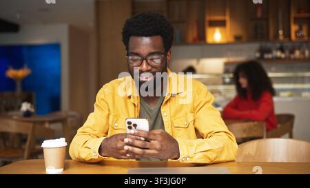 Lächelnder afroamerikanischer Mann mit Bart und Brille, der in einem Café auf einem Smartphone tippt. Mann sitzt an einem Holztisch mit Kaffeetasse und Laptop im Innenraum Stockfoto