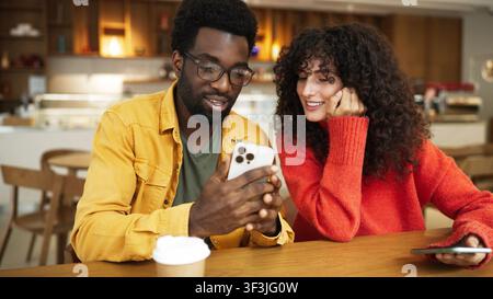 Mann in gelbem Hemd zeigt Smartphone einer Frau in rotem Pullover am Cafétisch. Afroamerikanischer Mann mit Brille hält Telefon, während lockighaarige Stockfoto