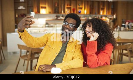 Lächelnder afroamerikanischer Mann in gelbem Hemd und lockige kaukasische Frau in rotem Pullover, die zusammen im Café ein Selfie machen. Glückliches Paar, das posiert Stockfoto