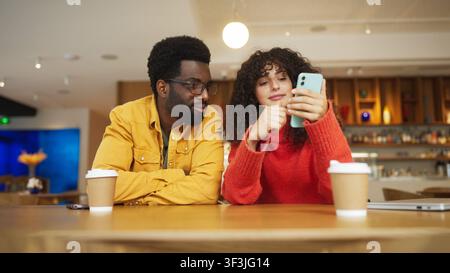Mann mit gemischter Herkunft in gelbem Hemd und Frau in rotem Pullover sitzen an einem Café-Tisch mit Kaffeetassen und schauen gemeinsam auf ein Smartphone. Mann mit Brille Stockfoto