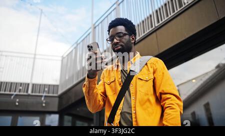 Attraktiver afroamerikanischer Mann mit Bart trägt Brille und hält ein Smartphone im Freien. Mann mit Umhängetasche steht in der Nähe eines modernen Gebäudes und lächelt Stockfoto
