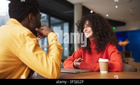 Kaukasische Frau mit lockigem Haar lächelt und spricht mit einem afroamerikanischen Mann drinnen in einem Café. Freunde sitzen an einem Holztisch mit einer Kaffeetasse und Stockfoto