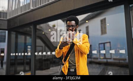 Attraktiver afroamerikanischer Mann mit Bart, der eine Brille trägt und ein Smartphone im Freien hält, überrascht schaut. Mann, der emotional auf gute Nachrichten reagiert oder Stockfoto