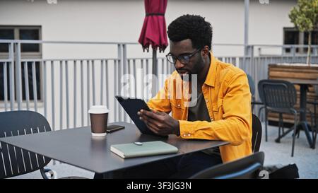 Attraktiver afroamerikanischer Mann mit Bart, der draußen an einem Tisch ein digitales Tablet benutzt. Mann mit Brille, der sich auf den Bildschirm konzentriert, mit Kaffeetasse und Notizbuch Stockfoto