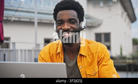 Porträt eines gutaussehenden afroamerikanischen Mannes mit Bart, der ein Headset trägt und lächelnd im Freien an einem Laptop arbeitet. Mann genießt Fernkommunikation Stockfoto
