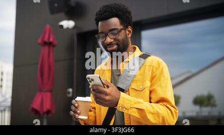 Ein lächelnder Mann mit Brille und gelbem Hemd überprüft sein Handy, während er eine Kaffeetasse vor einem modernen Gebäude hält Stockfoto