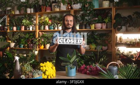 Porträt eines schönen professionellen Floristen, der in einem Blumenladen mit geschlossenem Schild steht, eine Schürze und eine Brille trägt. Erfolgreicher Mannarbeiter schaut Stockfoto