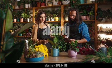Zwei attraktive erfolgreiche Mitarbeiter der Blumensphäre, die Pflanzen in einem Blumenladen beaufsichtigen. Junge schöne Frau und gutaussehender langhaariger Mann Stockfoto