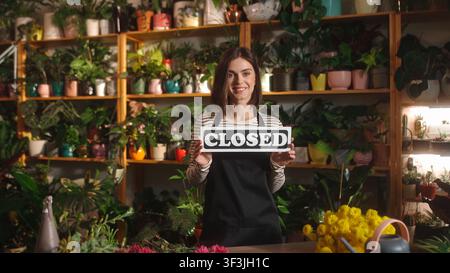 Porträt einer atemberaubend attraktiven professionellen Floristin, die im Blumenladen steht und ein „Geschlossen“-Schild trägt und eine Schürze trägt. Erfolgreiche Arbeiterin schaut Stockfoto