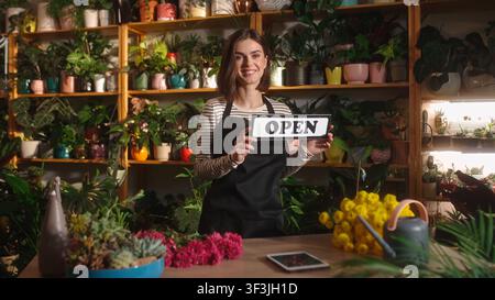 Porträt einer niedlichen hübschen kaukasischen Frau, die ein offenes Schild hält und zeigt, eine Schürze trägt und in einem Blumenladen arbeitet. Attraktive junge Floristin im Stockfoto