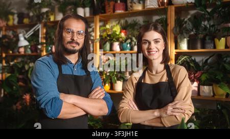 Portrait von zwei jungen, schönen professionellen Floristen, die im Blumenladen arbeiten, in die Kamera blicken, lächeln und mit verschränkten Armen posieren. Selbstbewusste Langhaarige Stockfoto
