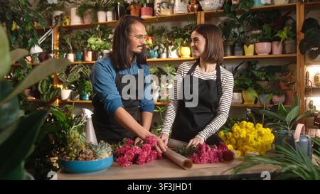 Zwei professionelle schöne junge Floristen dekorieren einen Blumenstrauß mit einer Verpackung auf der Theke im Blumenladen. Süße Frau und gutaussehender Mann Stockfoto