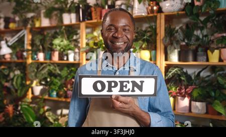 Glücklicher erfolgreicher afroamerikanischer junger Mann hält ein offenes Schild und schaut lächelnd in die Kamera. Schöner bärtiger Erwachsener arbeitet als Florist in einem gemütlichen Stockfoto