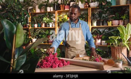 Fröhlicher professioneller Florist, der einen schönen Blumenstrauß in einem gemütlichen Blumenladen macht und dekoriert, schaut in die Kamera und lächelt. Schöne Afrikanerin Stockfoto