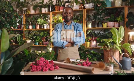 Fröhlicher professioneller Florist, der einen schönen Blumenstrauß in einem gemütlichen Blumenladen macht und dekoriert, schaut in die Kamera und lächelt. Schöne Afrikanerin Stockfoto