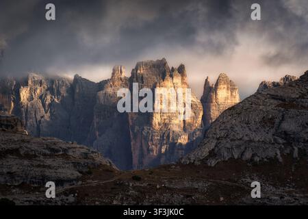Dramatische alpine Landschaft des schroffen Berggipfels Tre Cime, der in warmem goldenem Sonnenlicht eines Sonnenuntergangs vor dunklen stürmischen Wolken in den italienischen Alpen, Dolomiten, S Stockfoto