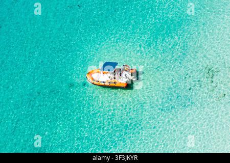 Luftbild-Drohnenaufnahme eines kleinen orangefarbenen Schlauchboots, das auf dem transparenten türkisfarbenen Wasser der Bucht Cala Macarella schwimmt, Menorca, Balearen, Spanien Stockfoto